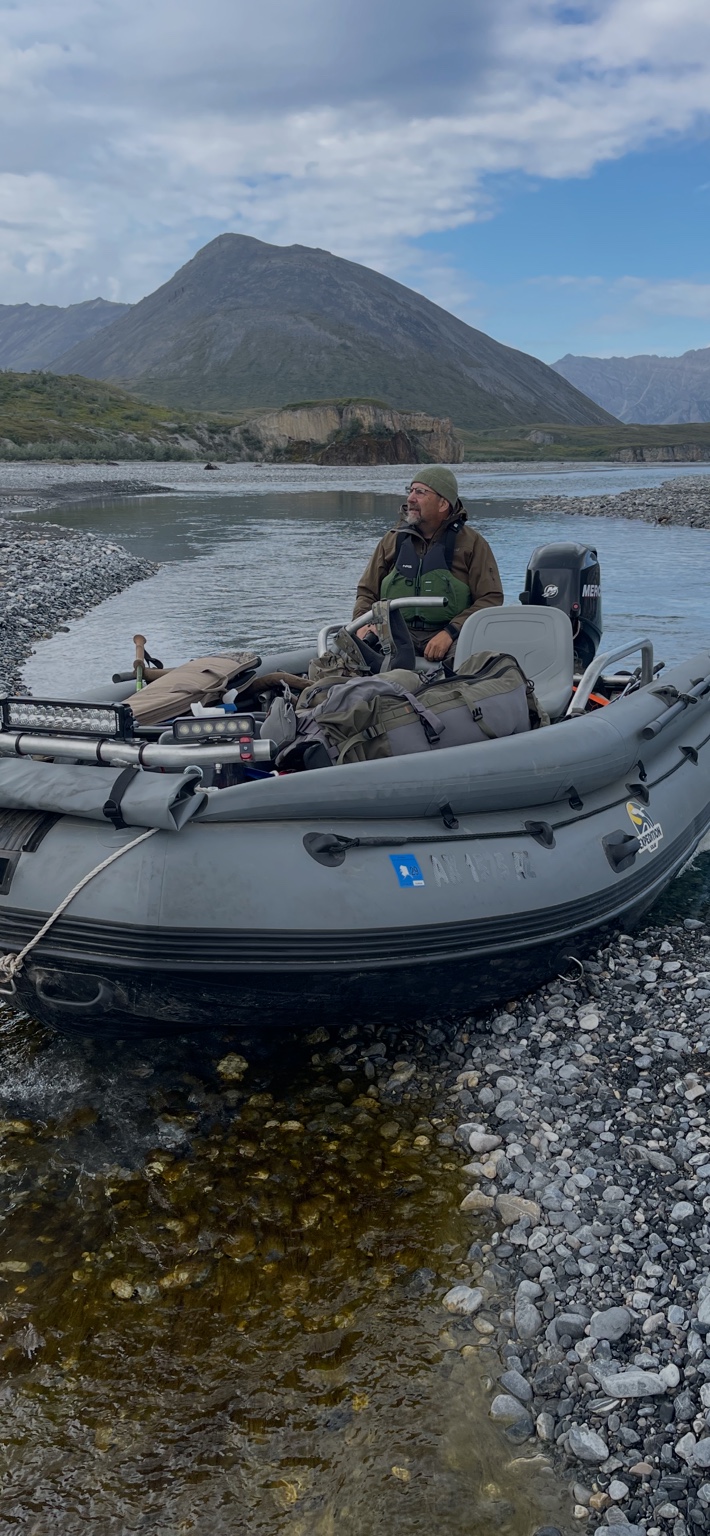 Tom Anderson seated in a custom-rigged gray inflatable riverboat on an Alaskan river with mountains behind
